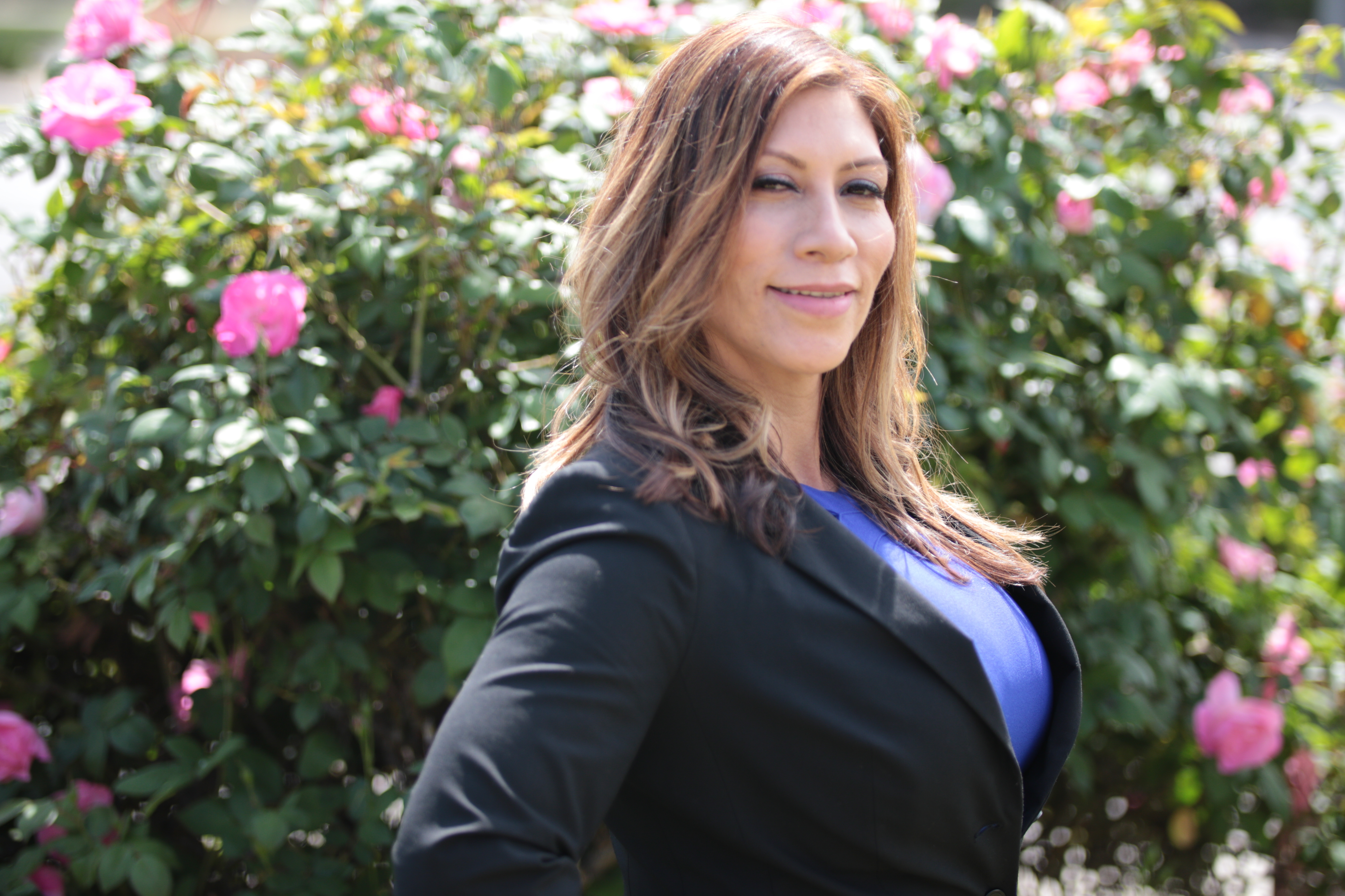 Maria Martinez in a black blazer and blue top standing in front of a pink rose bush
