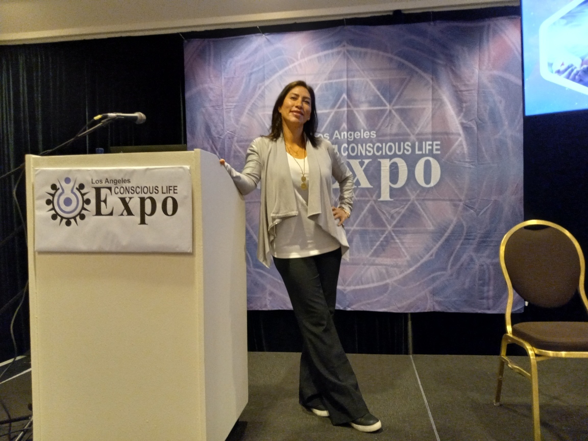 Maria Martinez standing at a podium in front of a Conscious Life Expo backdrop