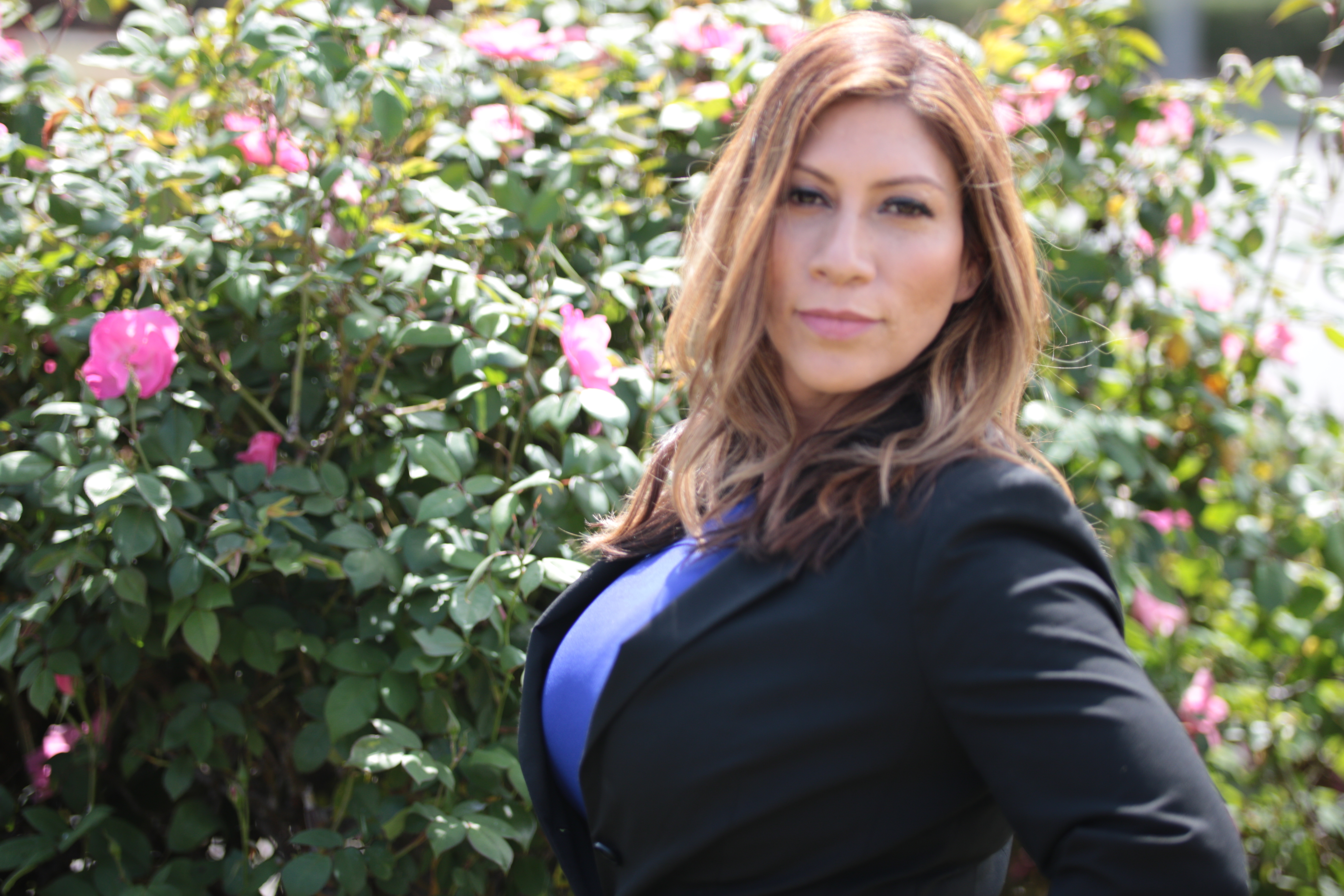 Maria Martinez in a black blazer and blue top beside a flowering pink rose bush